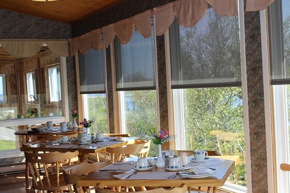 A dining area with multiple wooden tables and chairs, large windows with curtains, and a view of trees outside. - Duck Cove Inn (Margaree Harbour)