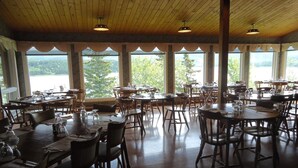 A restaurant with multiple tables and chairs, large windows with a view of trees, and wooden ceiling beams. - Duck Cove Inn (Margaree Harbour)