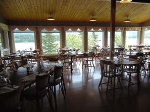 Un restaurante con varias mesas y sillas, amplias ventanas con vistas a árboles y vigas de madera en el techo.