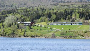 A lakeside lodge with a large building and a smaller structure, surrounded by dense forest and a hill in the background. - Duck Cove Inn (Margaree Harbour)
