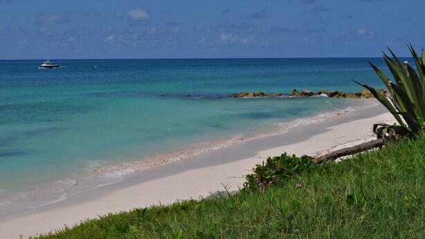 A beach with clear turquoise water, a boat in the distance, and a rocky outcrop with vegetation. - Sea View Apartment (Silver Sands)
