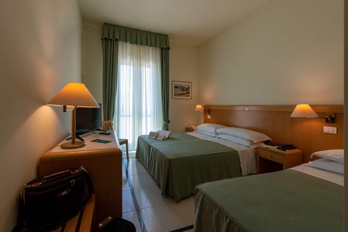 Quarto de hotel com duas camas, uma escrivaninha com luminária, uma cadeira, uma mala e uma janela com cortinas.