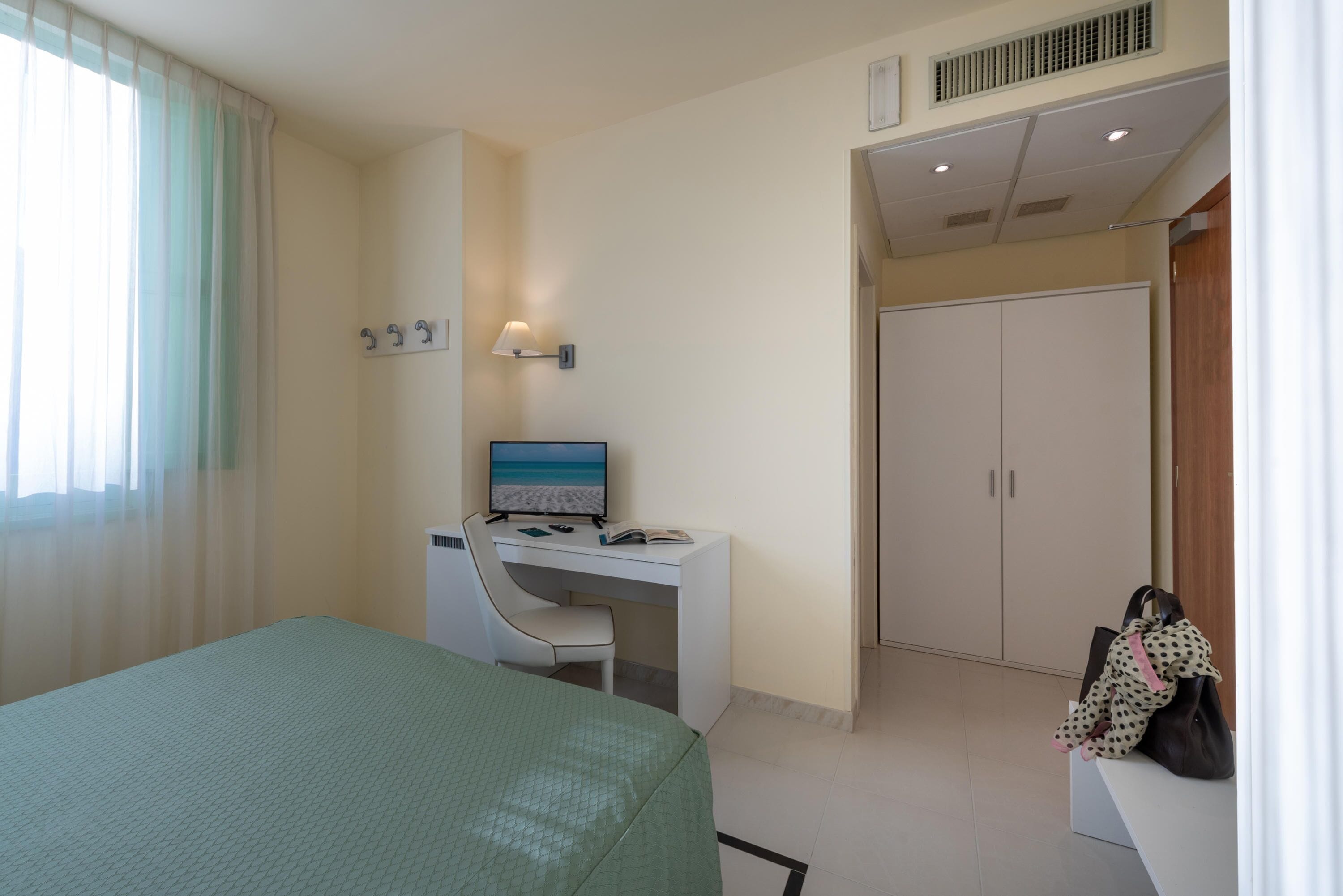 A hotel room with a bed, a desk with a computer, a chair, a wardrobe, and a window with curtains.