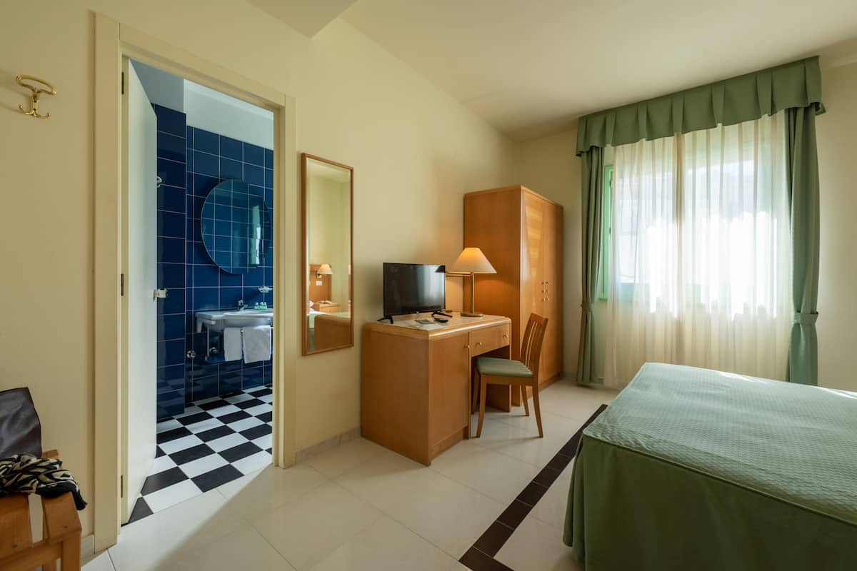 Quarto de hotel com chão quadriculado, uma escrivaninha com cadeira, cama, televisão e banheiro com espelho e pia.