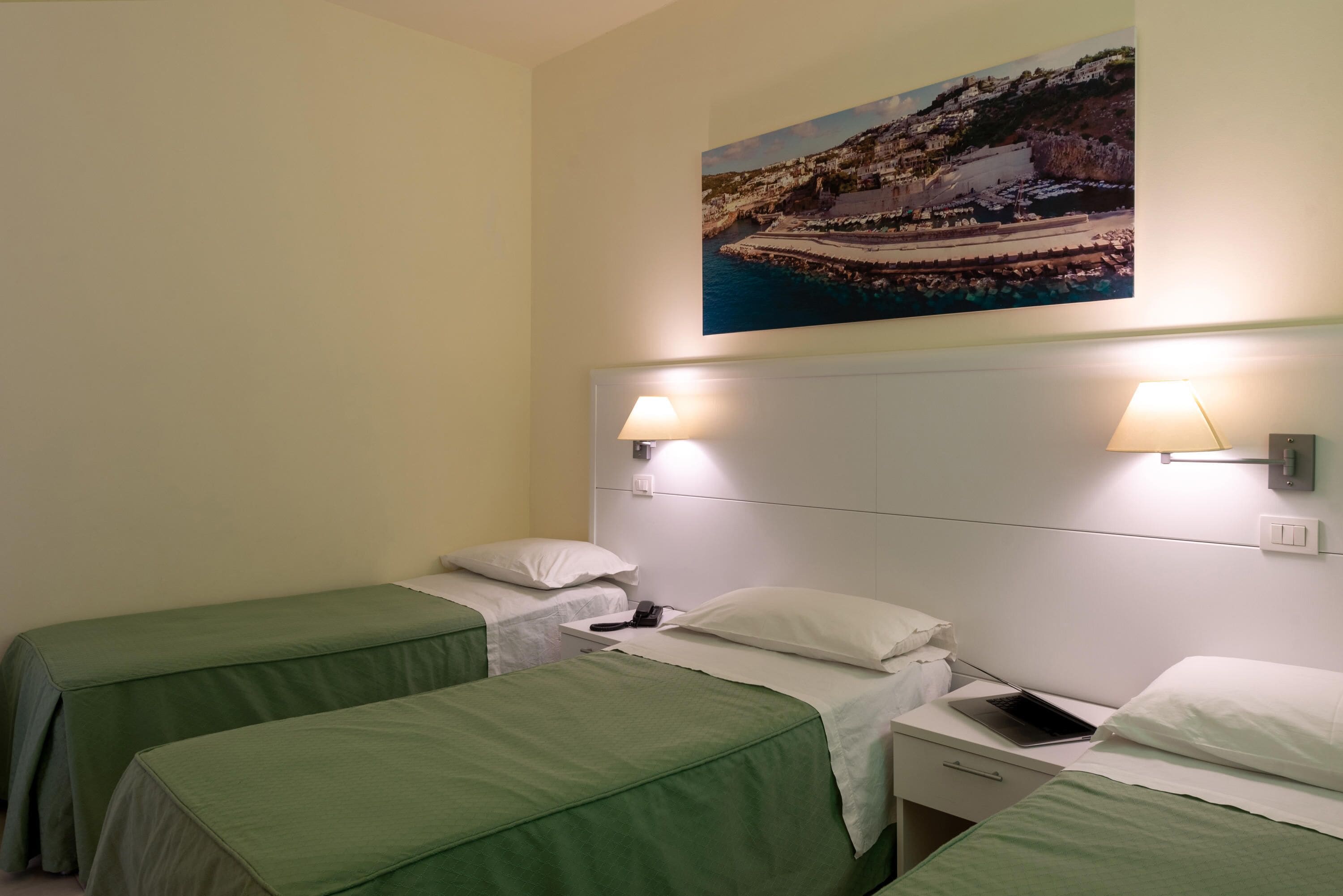 A hotel room with two single beds, a nightstand with a lamp, a phone, and a laptop.