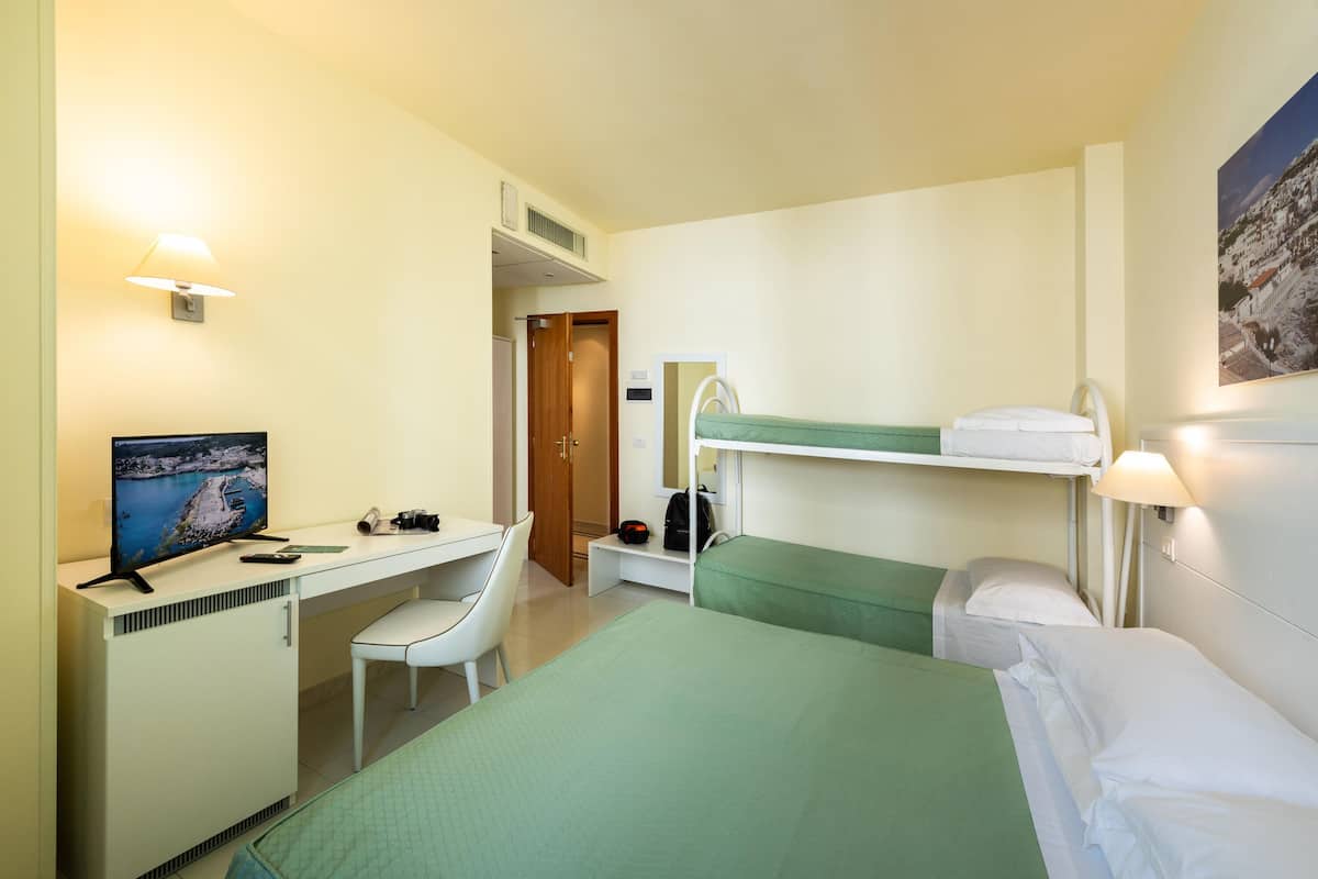 Quarto de hotel com uma cama, uma escrivaninha com computador, uma cadeira, uma televisão e uma beliche.