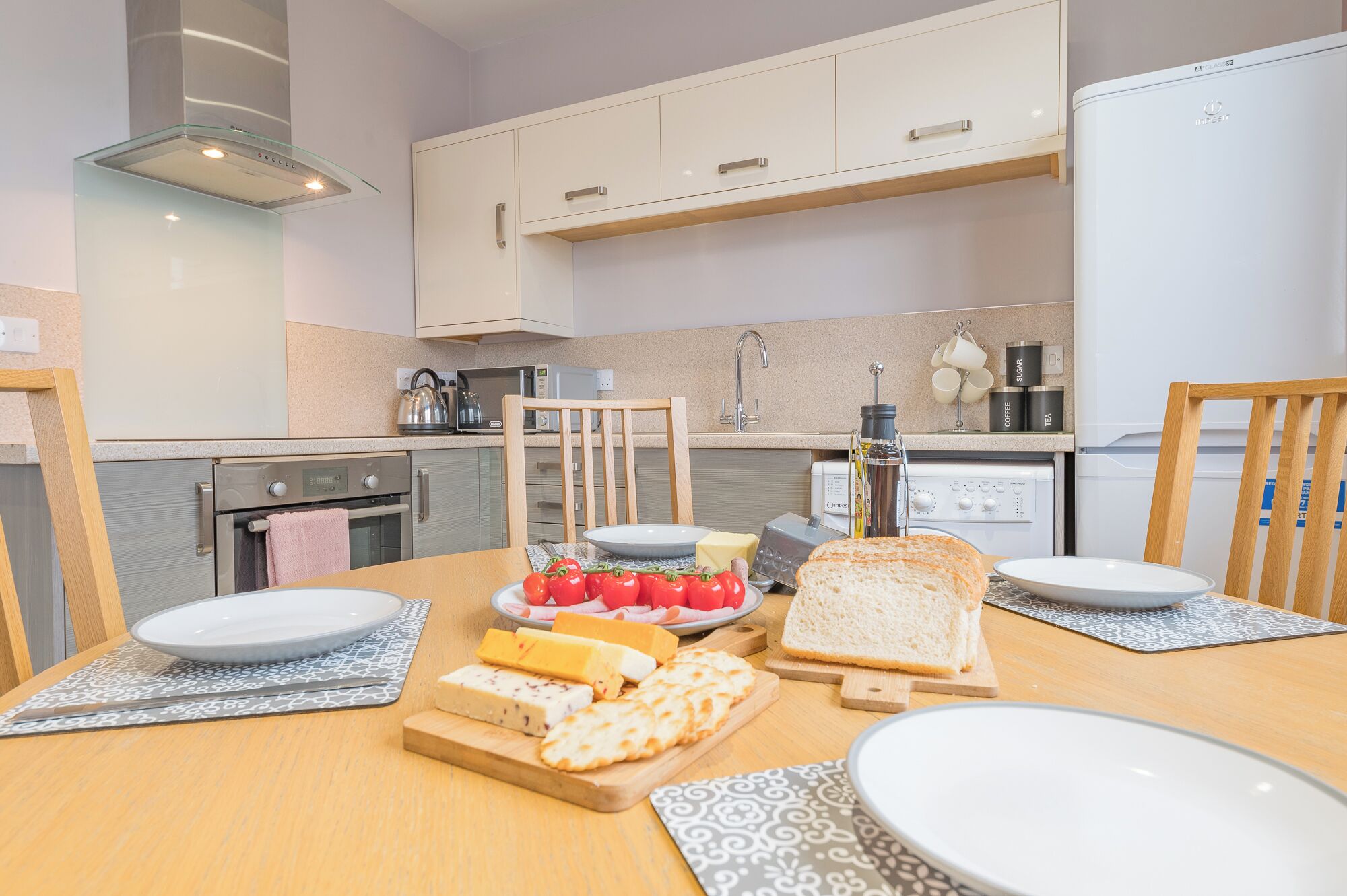 A modern kitchen with white cabinets, a built-in oven, and a microwave. There are two chairs, a dining table with plates, cheese, tomatoes, and bread.