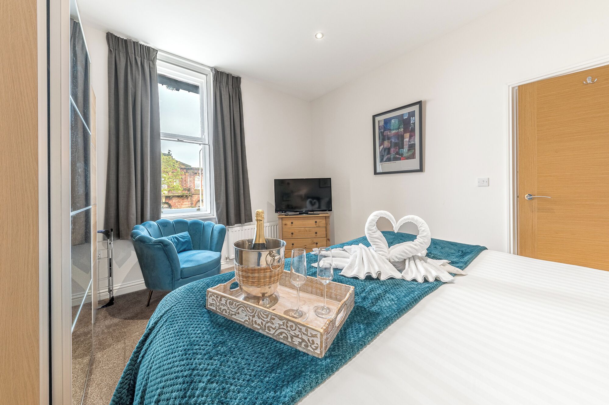 A hotel room with a bed, a blue armchair, a TV, a bottle of champagne, and a tray with glasses.