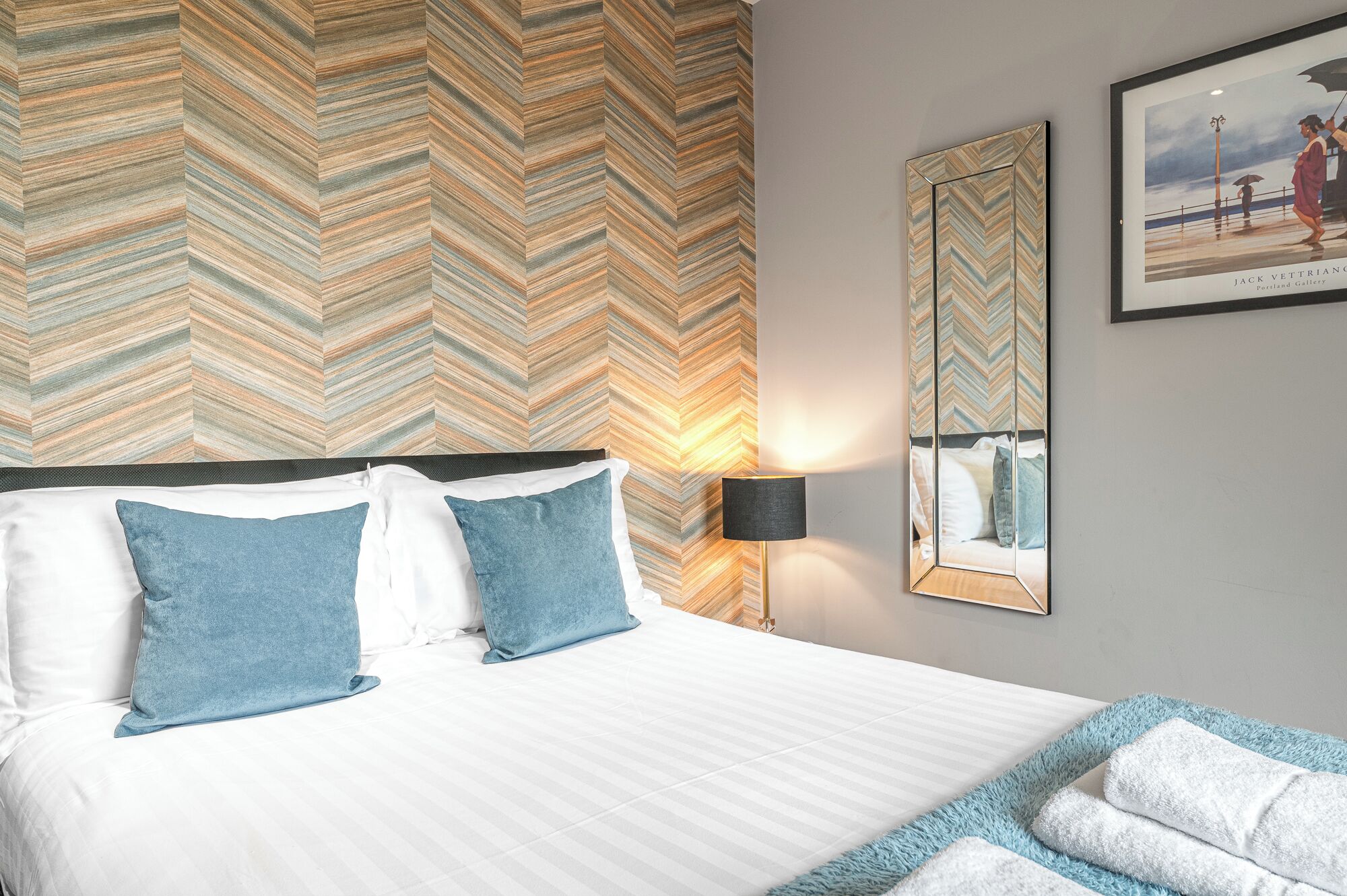 A hotel room with a bed, two pillows, a nightstand with a lamp, a mirror, and a framed picture on the wall.