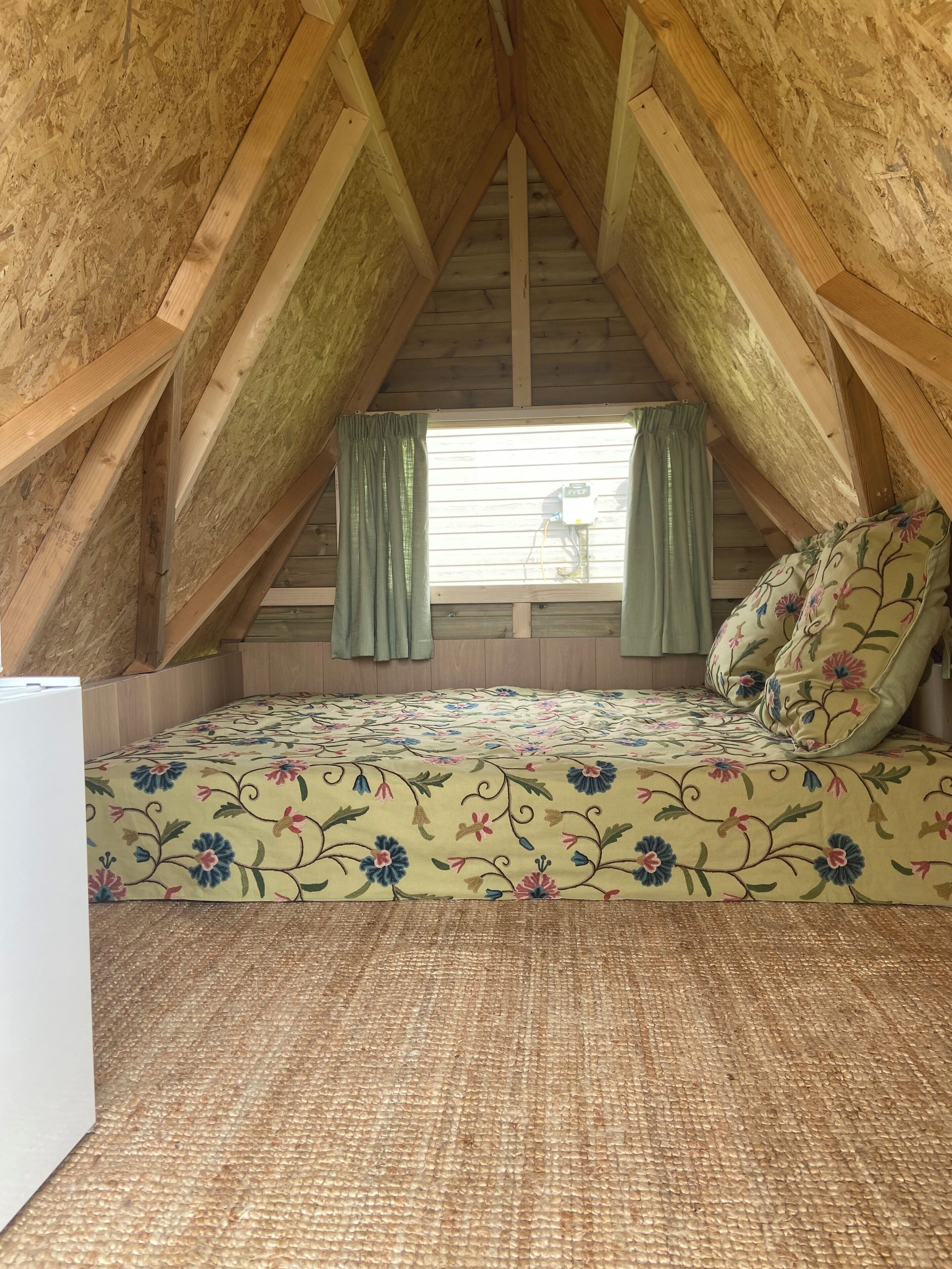 A small attic bedroom with a sloped ceiling, a bed with floral bedding, and a window with curtains.