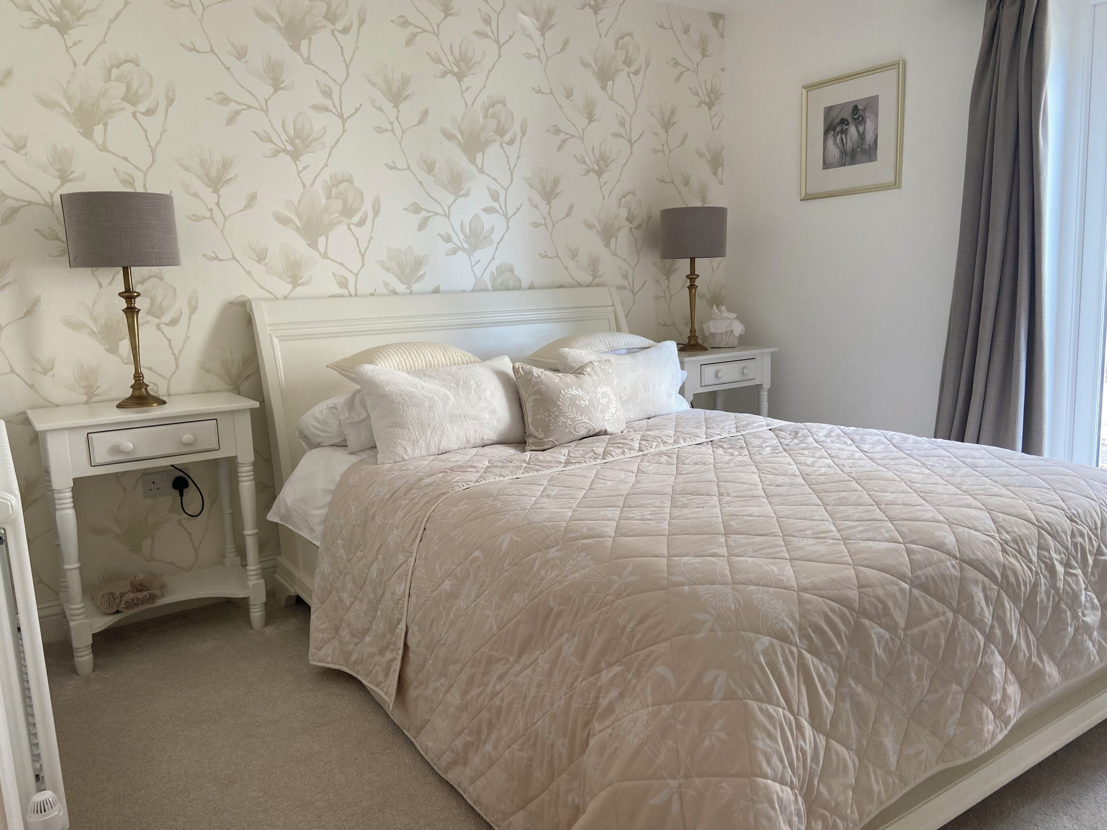 A neatly made bed with a quilted beige comforter, two bedside tables with lamps, a white nightstand, and a floral wallpaper pattern.