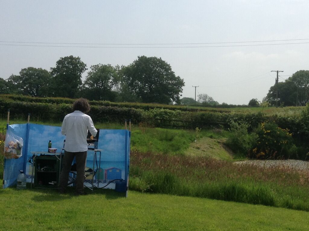 A person standing at a blue table with a laptop, surrounded by greenery and trees in the background.