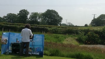 A person standing at a blue table with a laptop, surrounded by greenery and trees in the background.