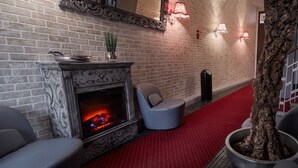 A room with a brick wall, a fireplace, two grey chairs, a red carpet, and a potted tree. - Pension Falconi (Kolin)