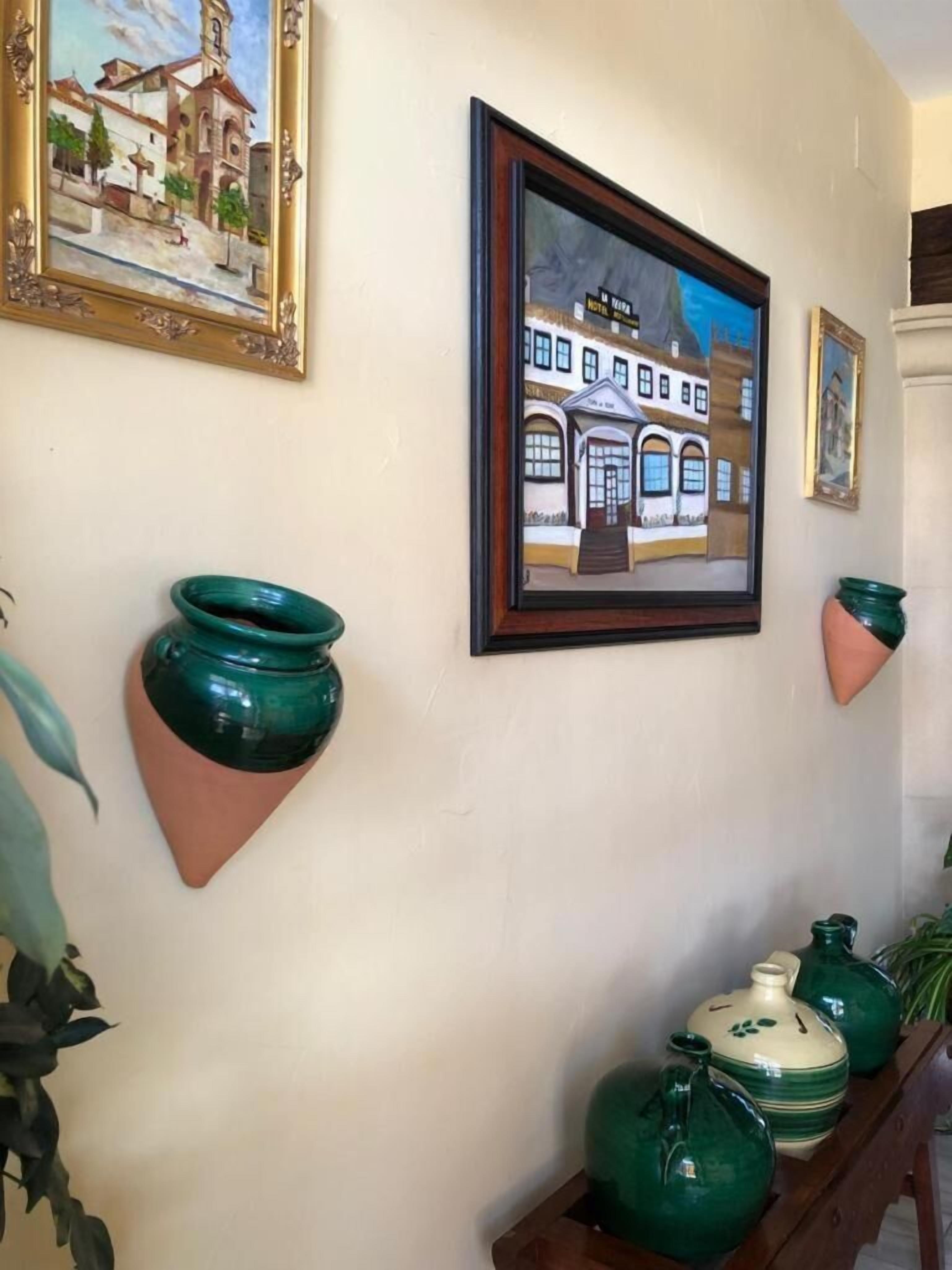 A wall with two framed paintings, a wooden shelf with green and beige ceramic pots, and a potted plant.