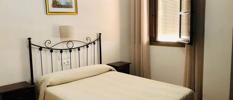 A bedroom with a bed, a nightstand, a lamp, a window with curtains, and a framed picture on the wall.