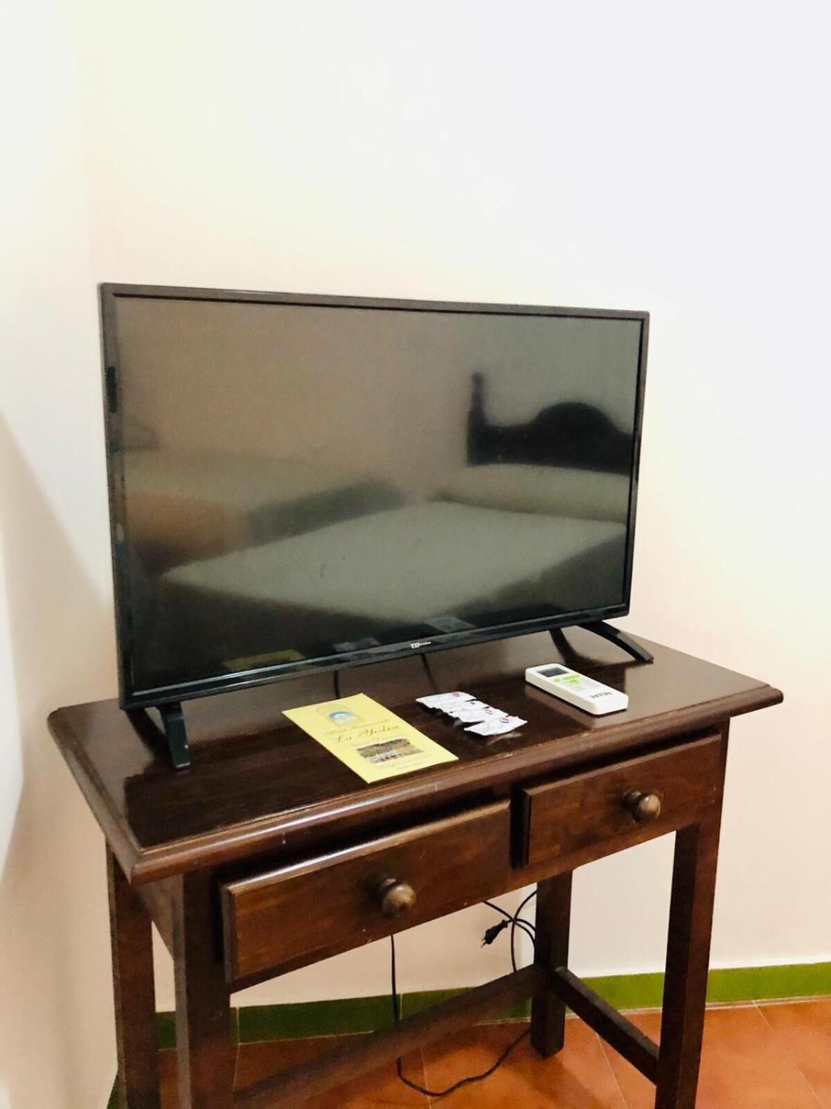 A television on a wooden stand with a bed in the reflection, a small drawer, and a power cord.