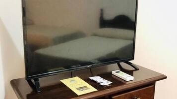 A television on a wooden stand with a bed in the reflection, a small drawer, and a power cord.