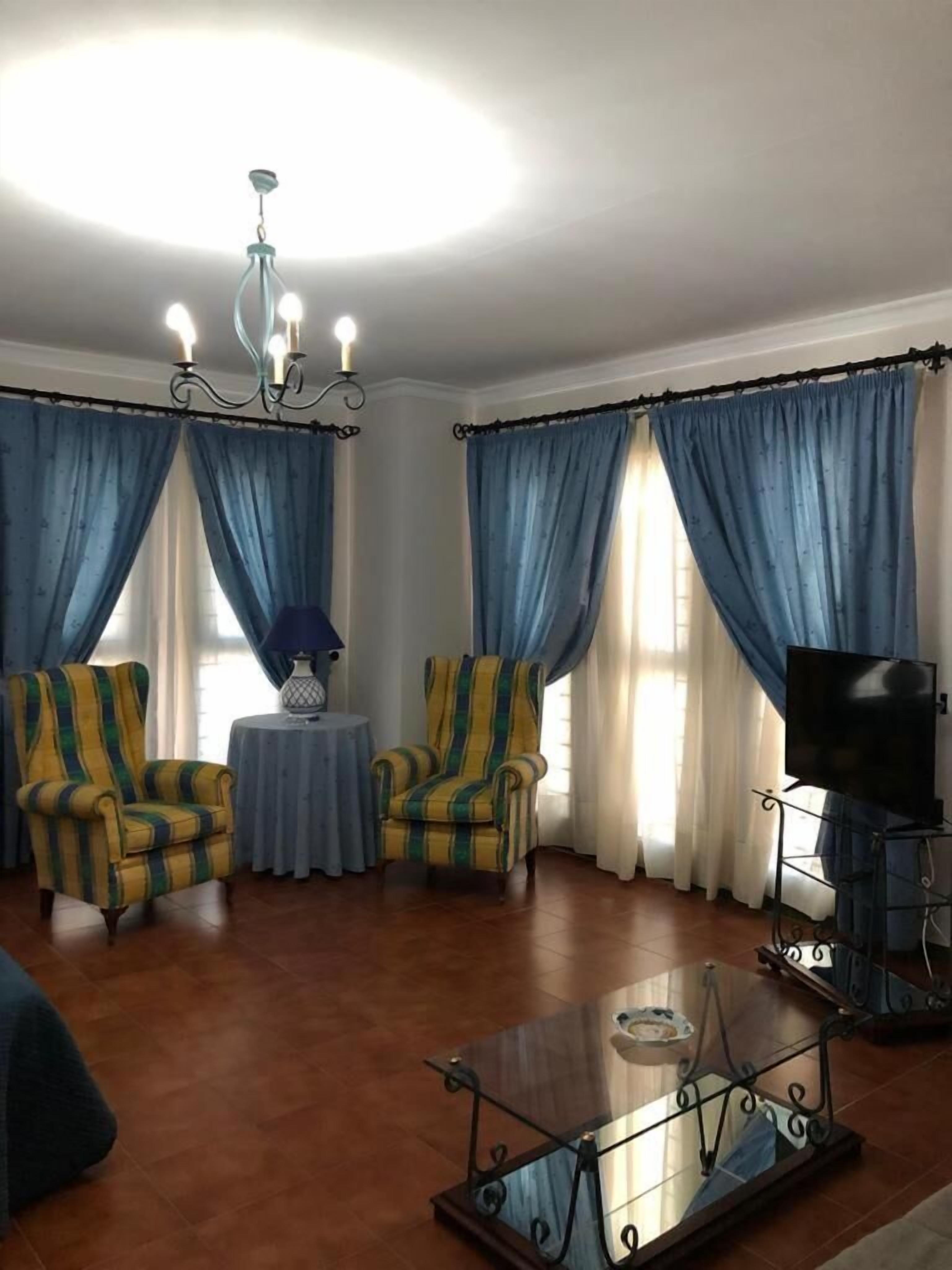 Una sala de estar con dos sillones, una mesa de centro de vidrio, un televisor sobre un mueble bajo y una araña.