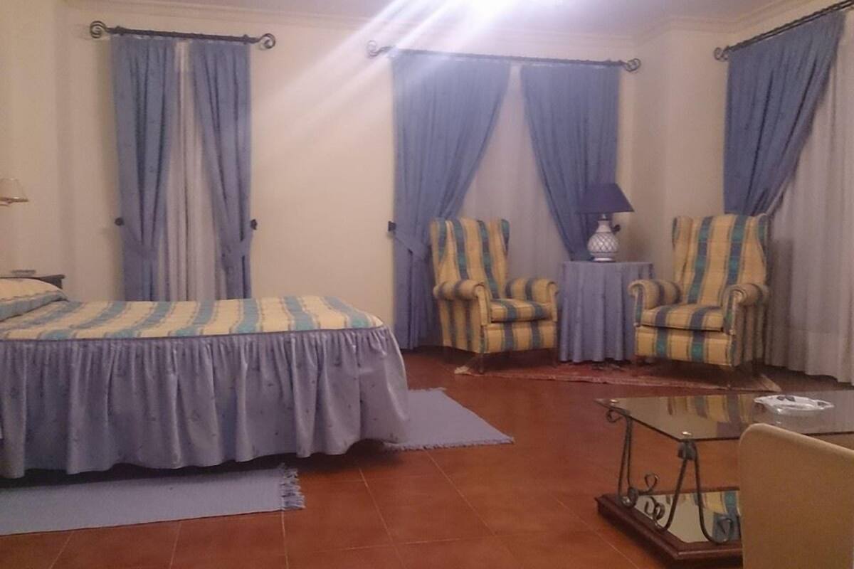 Un dormitorio con una cama, dos sillones, una mesita y una lámpara.