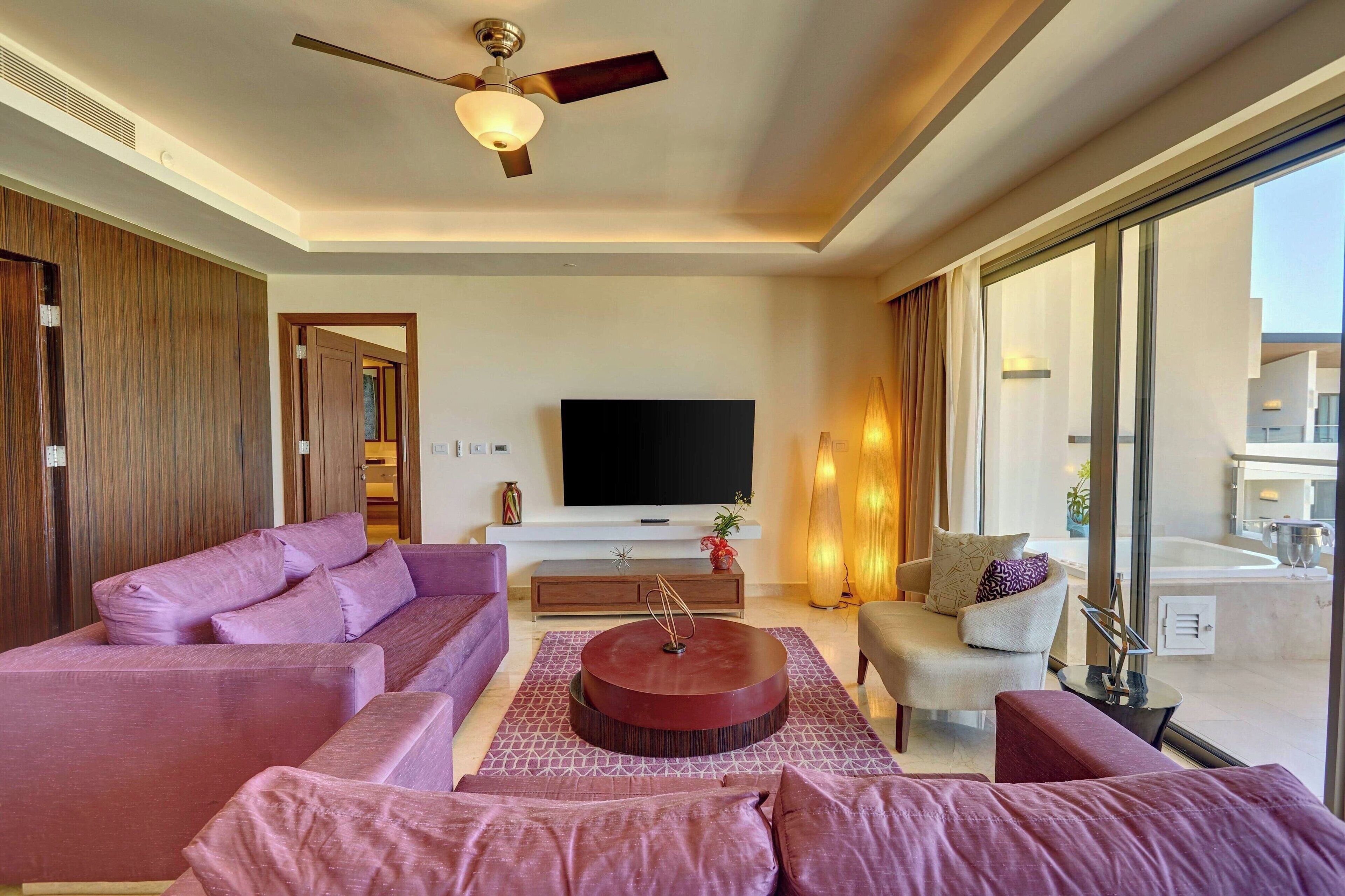 A modern living room with a large flat-screen TV, a ceiling fan, and a view of the pool and building outside.