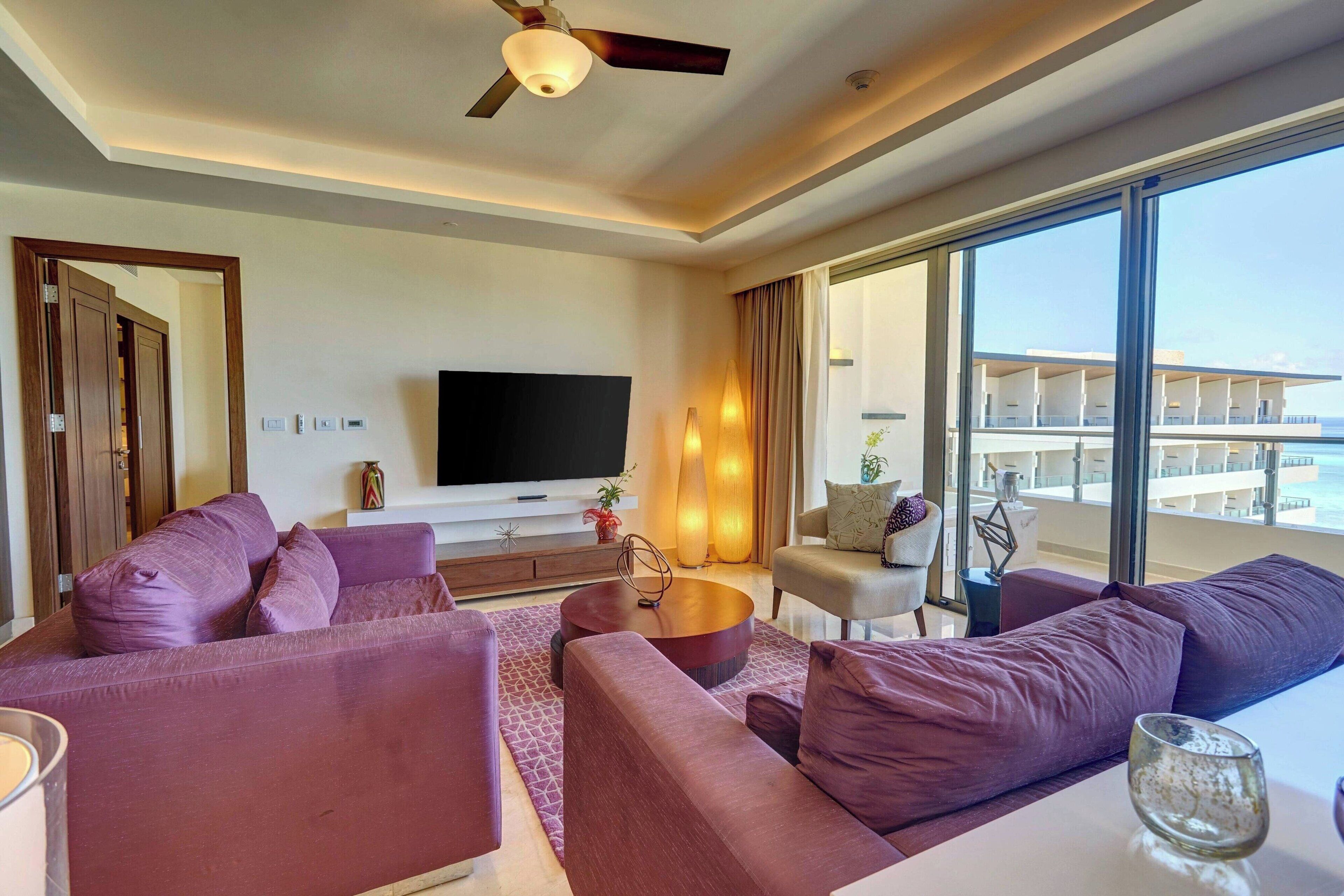 Una sala de estar moderna con un televisor de pantalla plana grande, un ventilador de techo y vistas a un balcón con mobiliario exterior.