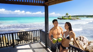 Une terrasse en bois, dotée d’une balustrade, surplombe une plage aux eaux turquoise et cristallines, ainsi qu’une petite île parsemée de palmiers.