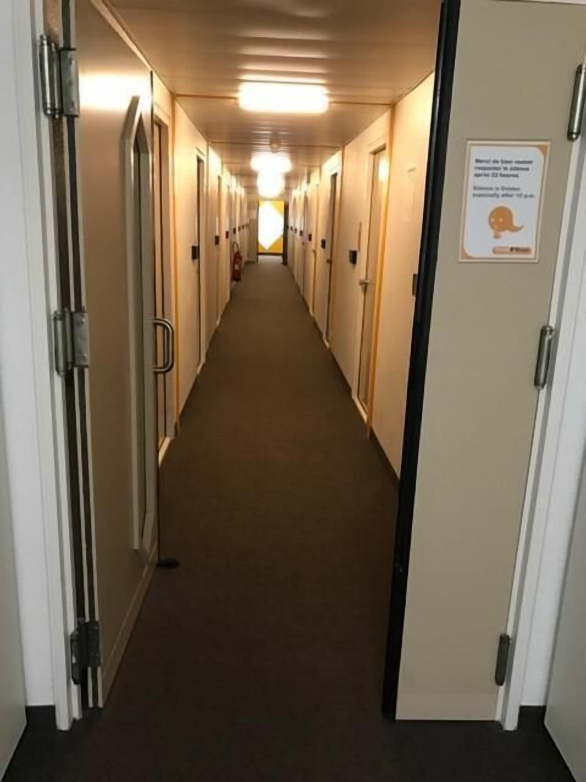 Un corridoio d'albergo con porte su entrambi i lati, pavimento rivestito di tappeto e una insegna sulla porta destra.