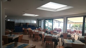 A spacious dining area with high ceilings, skylights, and large windows offering views of the outdoors. Tables are arranged for groups, with chairs and menus visible. - Hotel Ambra (Lignano Sabbiadoro)