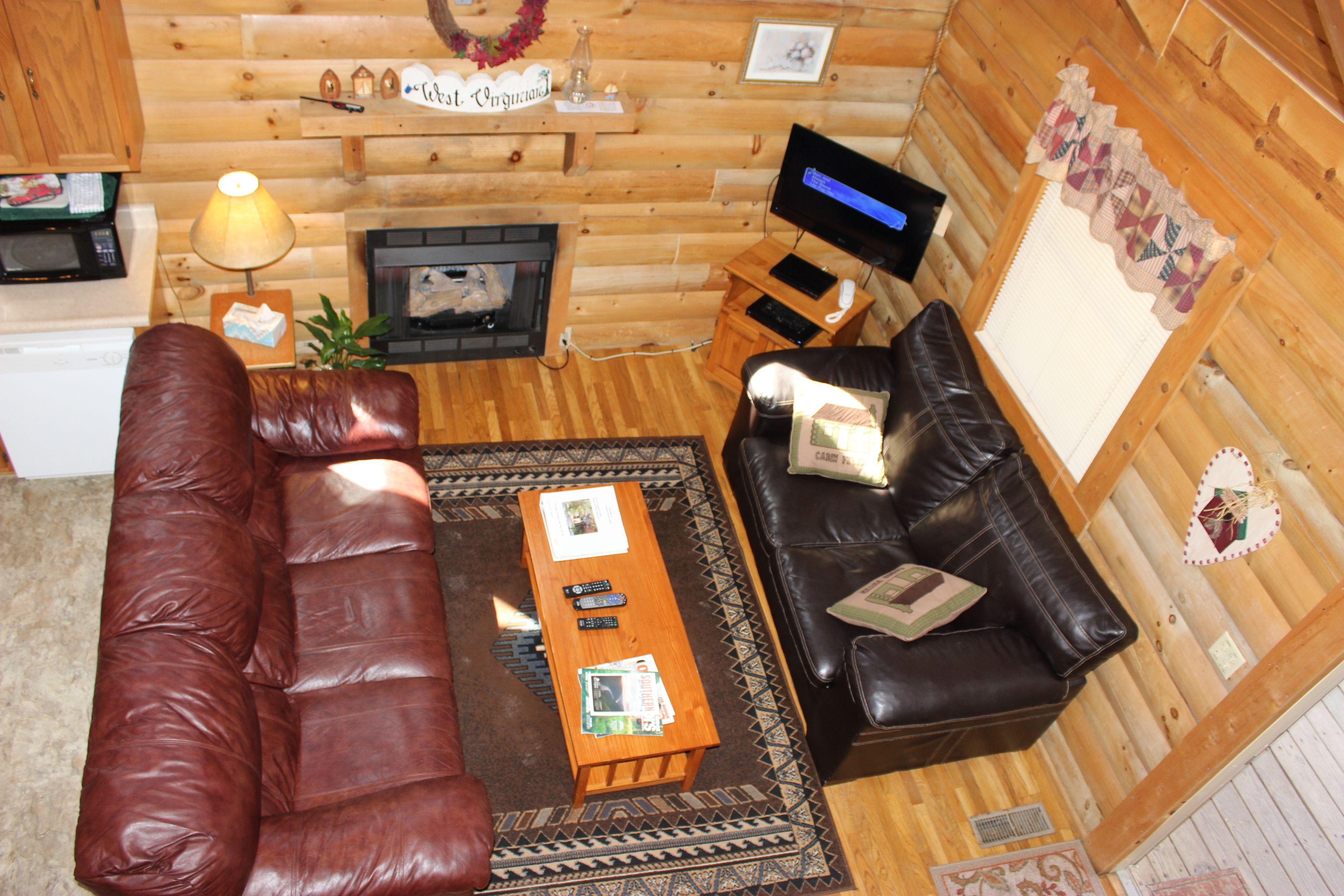 A cozy cabin interior with a brown leather couch, a smaller sofa, a wooden coffee table, a fireplace, a television, and a wooden wall with decorations.