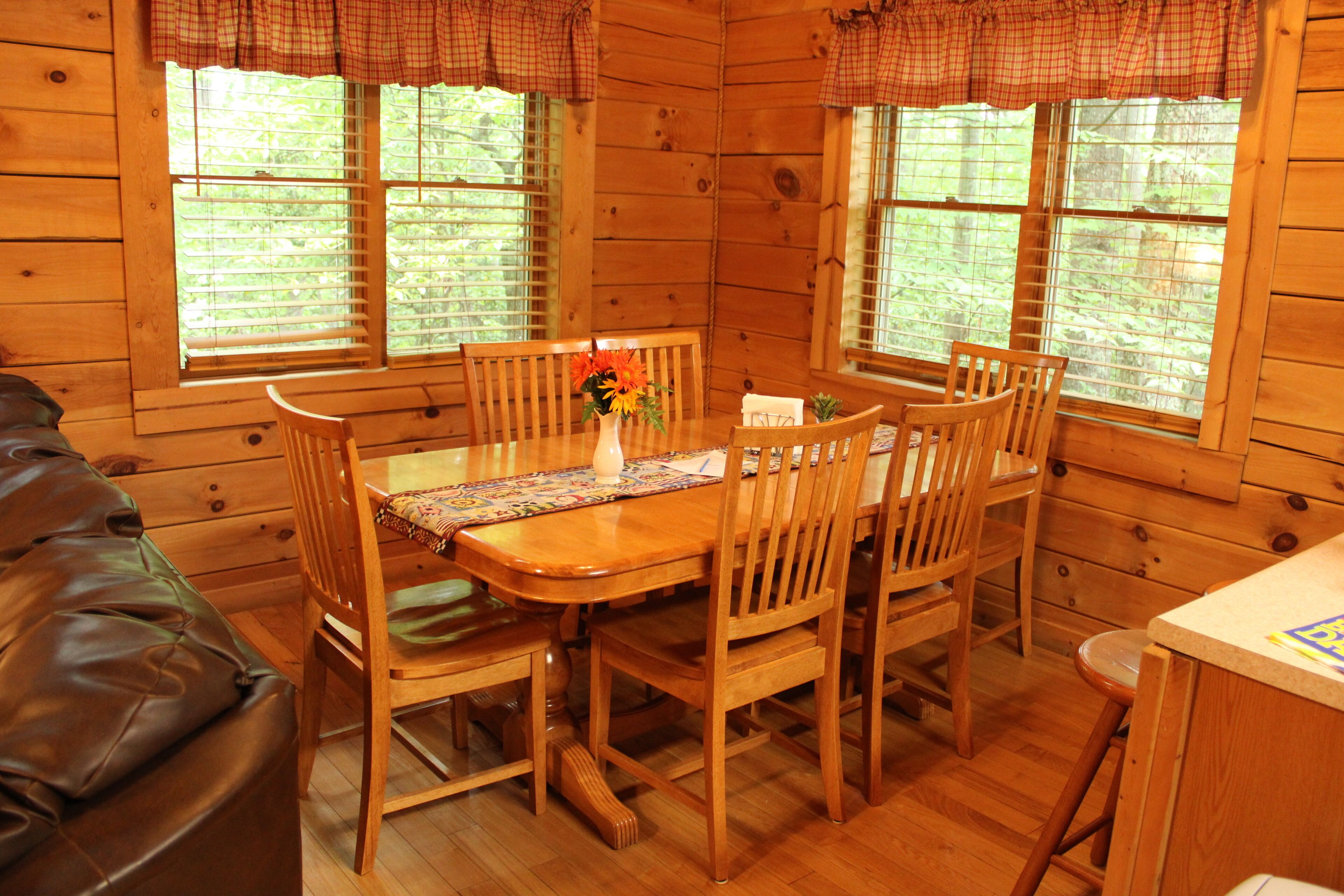 A wooden cabin interior with a dining table, chairs, a sofa, and large windows with blinds.