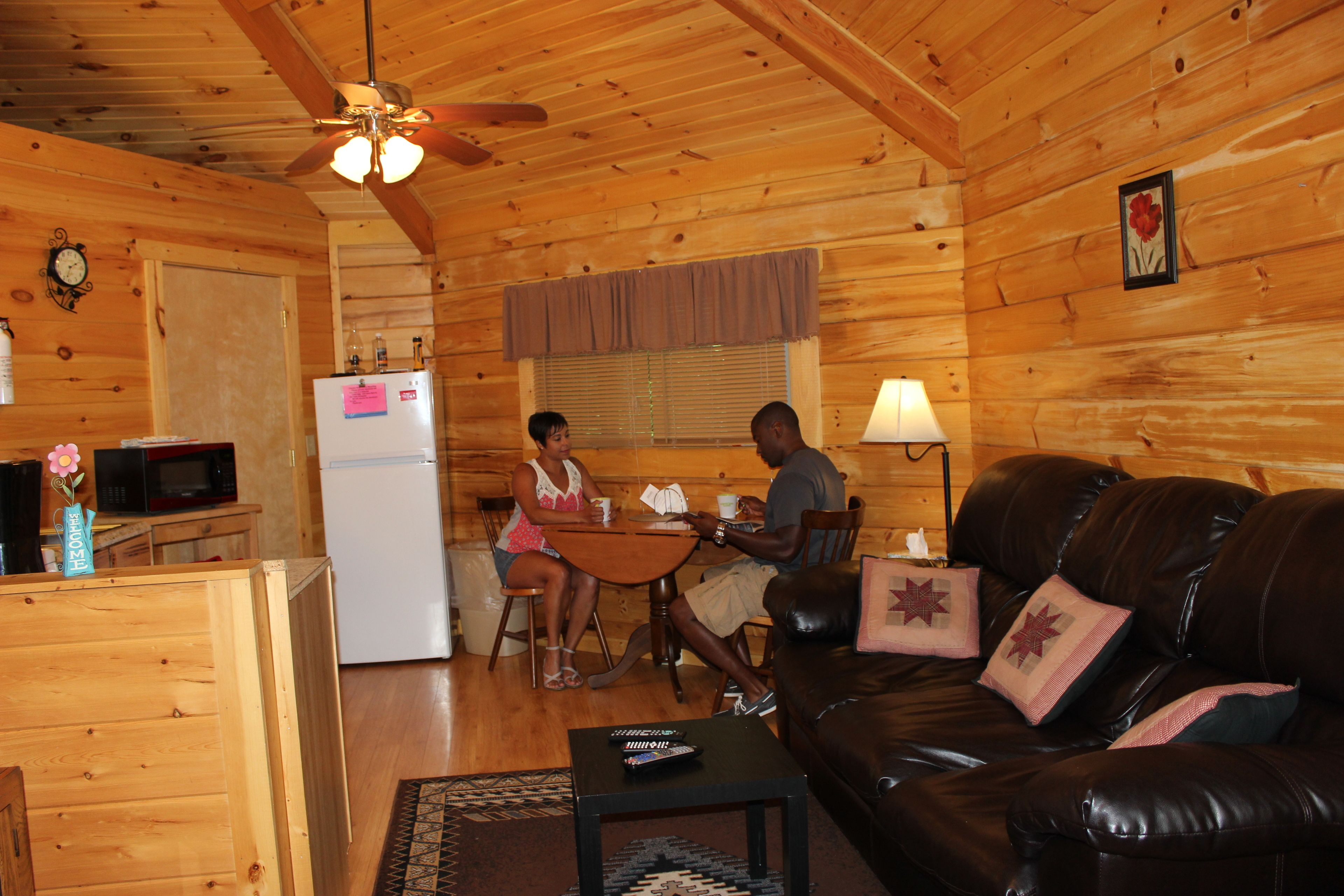 A wooden cabin interior with a dining area, a living room with a leather couch, and a kitchen with a refrigerator and microwave.