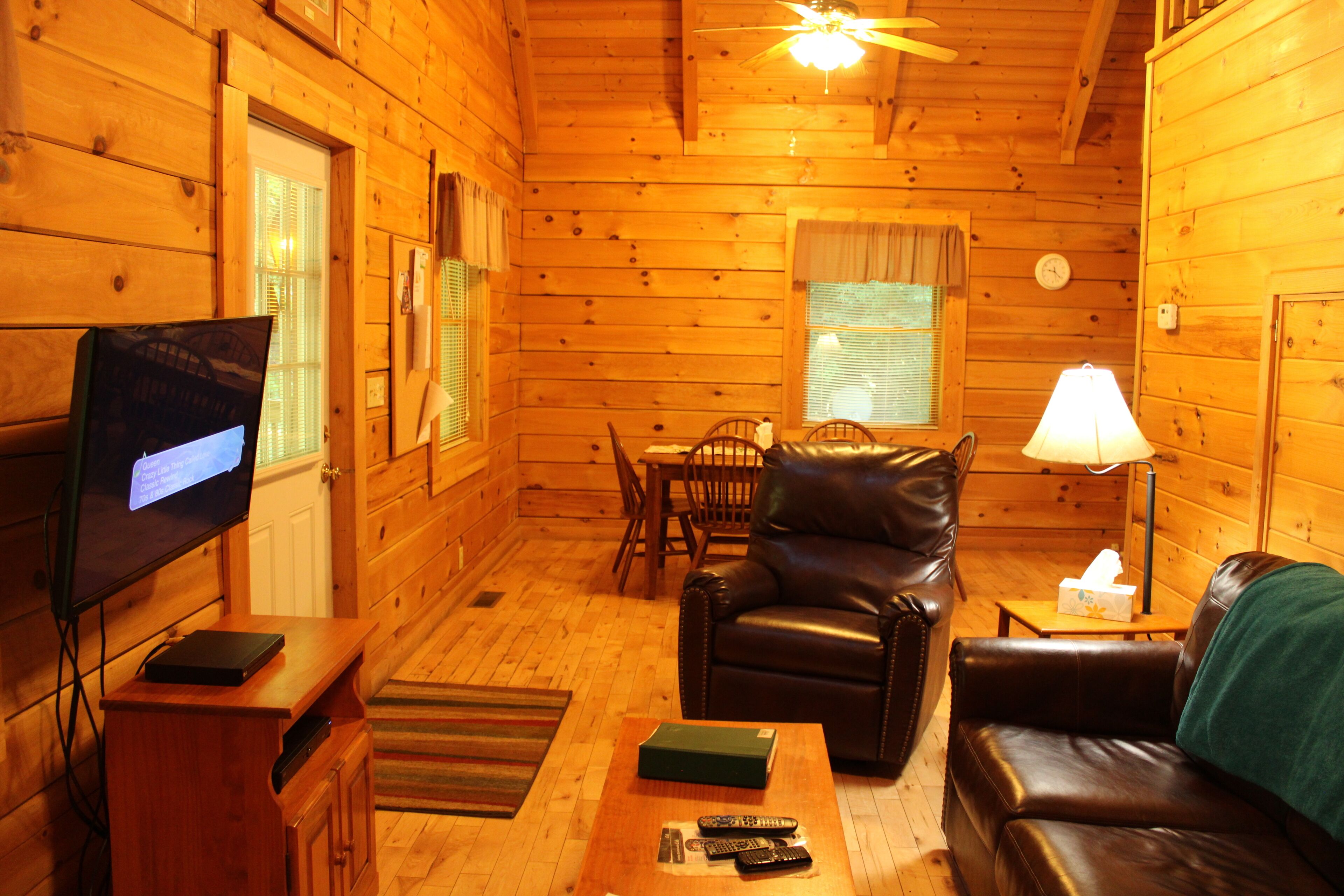 A cozy cabin interior with a leather sofa, a television, a dining table, and a lamp.