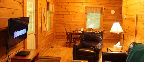 A cozy cabin interior with a leather sofa, a television, a dining table, and a lamp.
