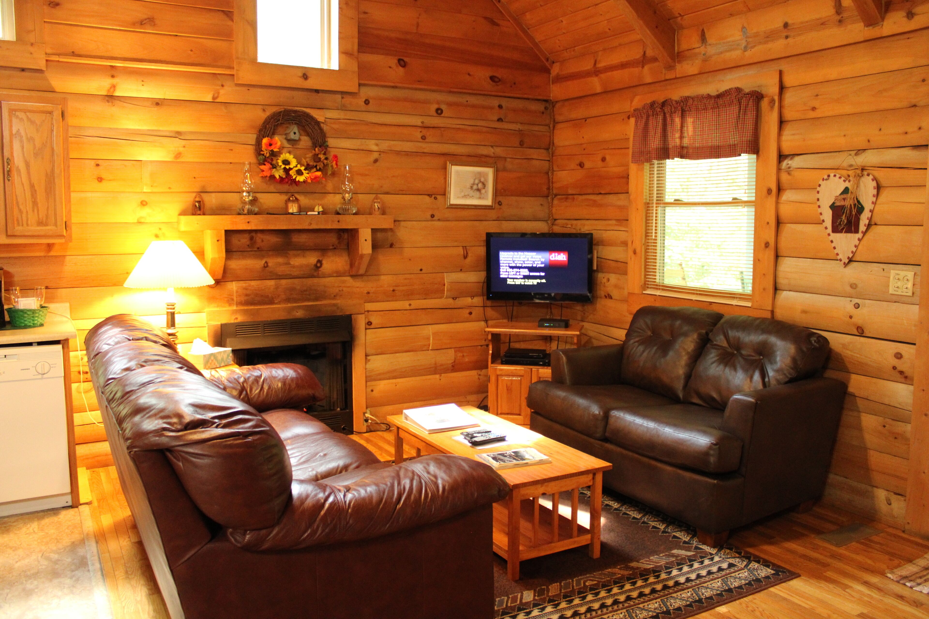 A cozy cabin interior with a brown leather sofa, a wooden coffee table, a television, and a fireplace.