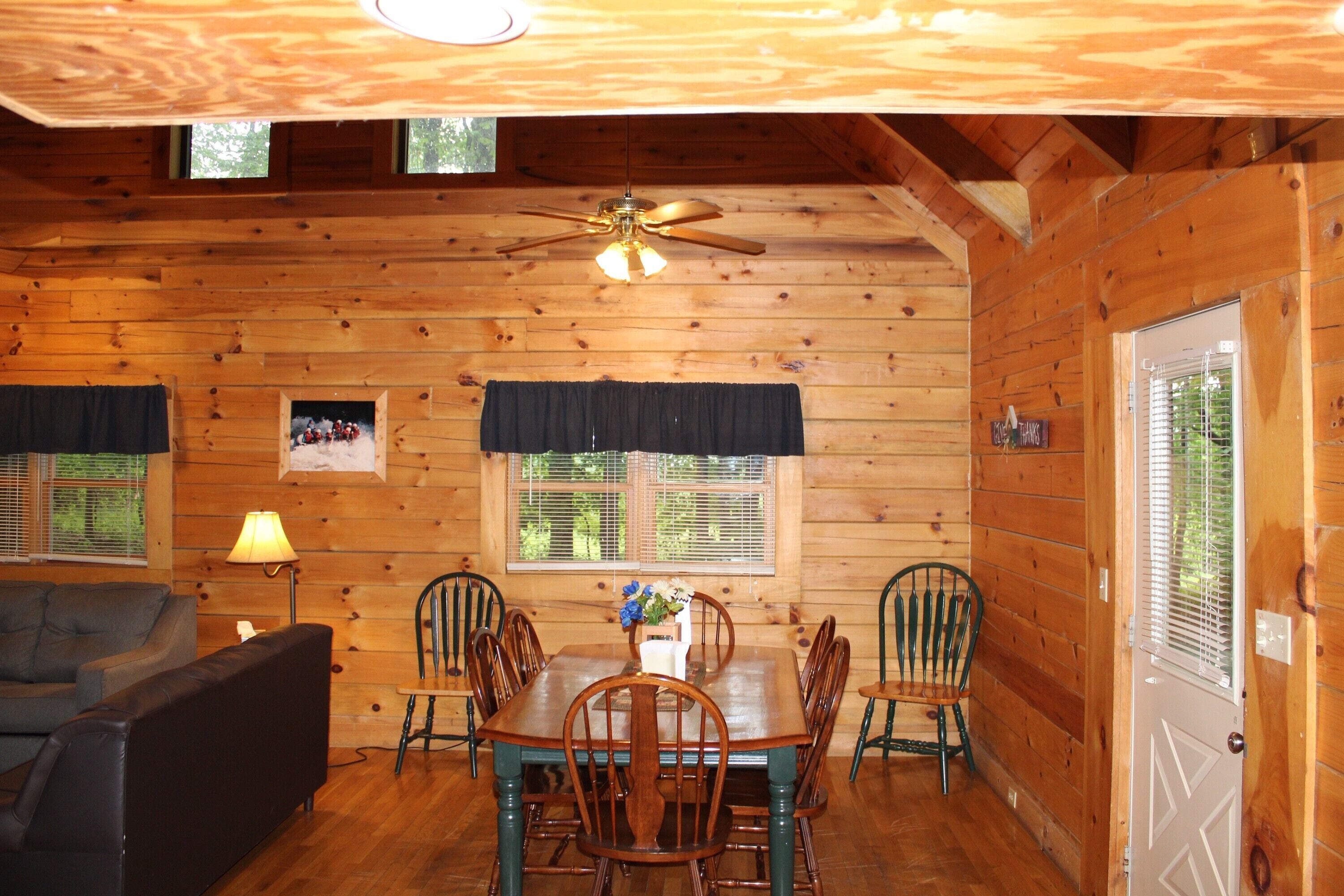 A wooden cabin interior with a dining table, chairs, a sofa, and a ceiling fan.