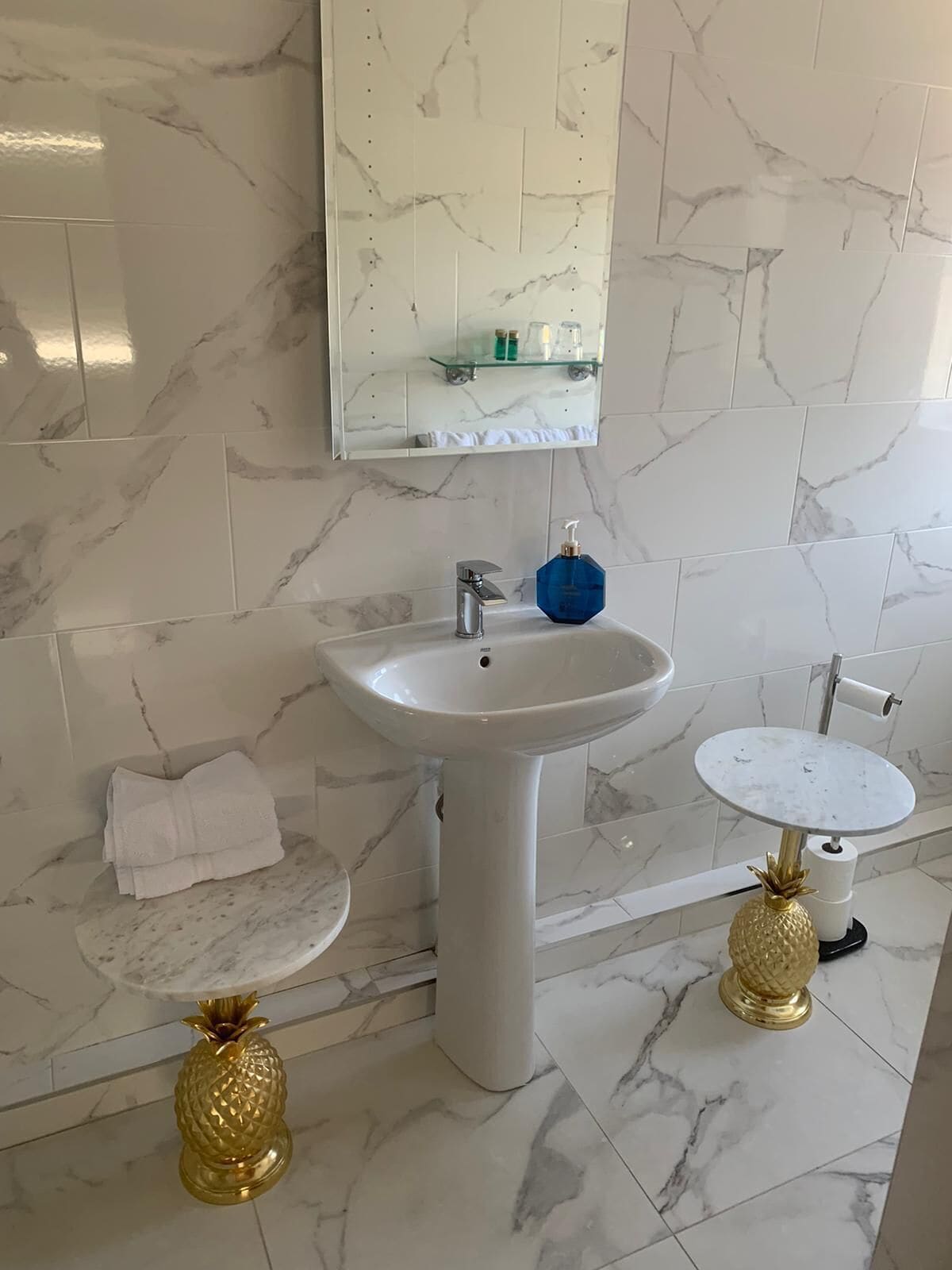 Un baño con lavamanos de mármol blanco, un lavamanos pequeño y redondo, y dos taburetes con forma de piña dorada.