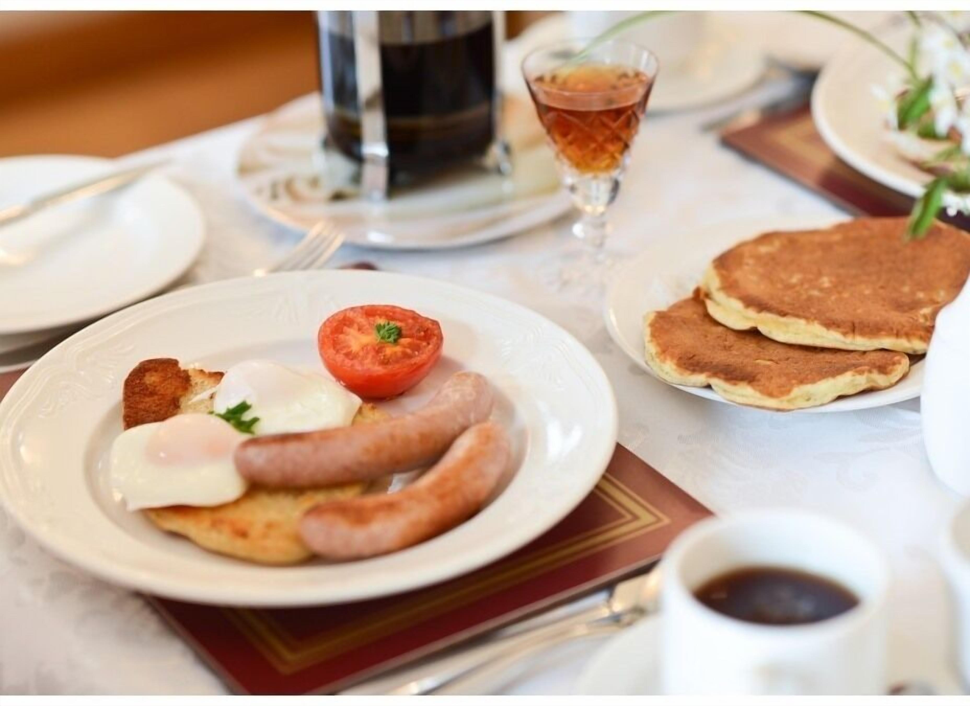 A breakfast setting with plates of food including sausages, a poached egg, and a tomato. There are pancakes on the side and a cup of coffee.