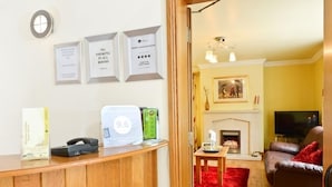 A reception desk with a 9.6 rating sign, framed certificates, and a no-smoking sign. A living room with a fireplace, a TV, and a red rug is visible through an open door. - Brooke Lodge Guesthouse (Magherafelt)