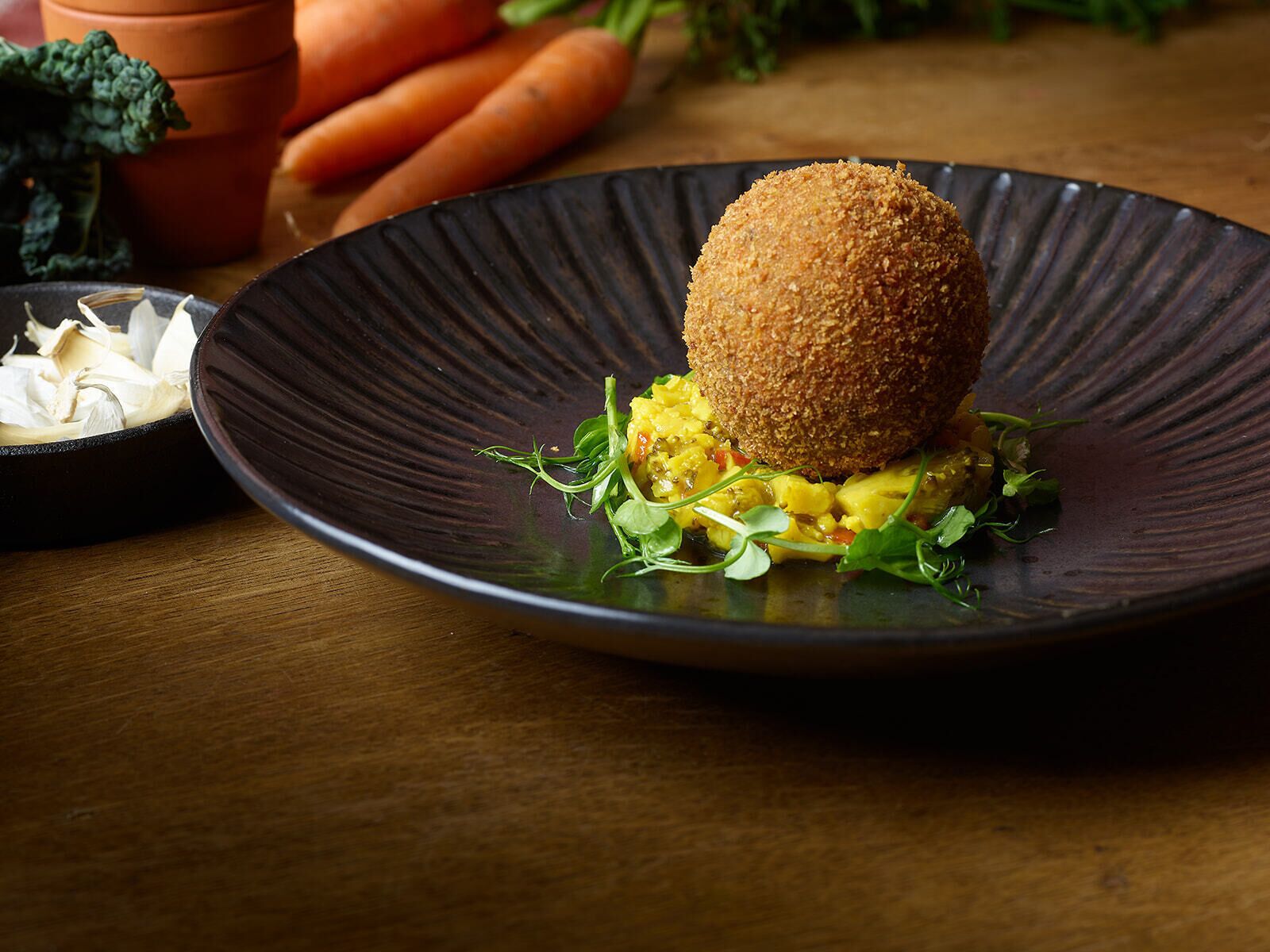 A dish with a breaded ball on top, served on a dark plate with a side of greens and a small bowl of garlic.