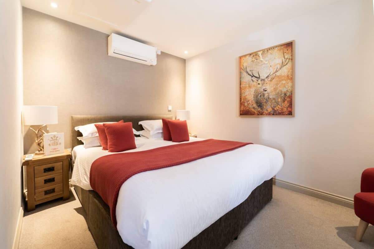 A neatly made bed with white linens and red accents, a bedside table with a lamp, and a wall-mounted air conditioner.