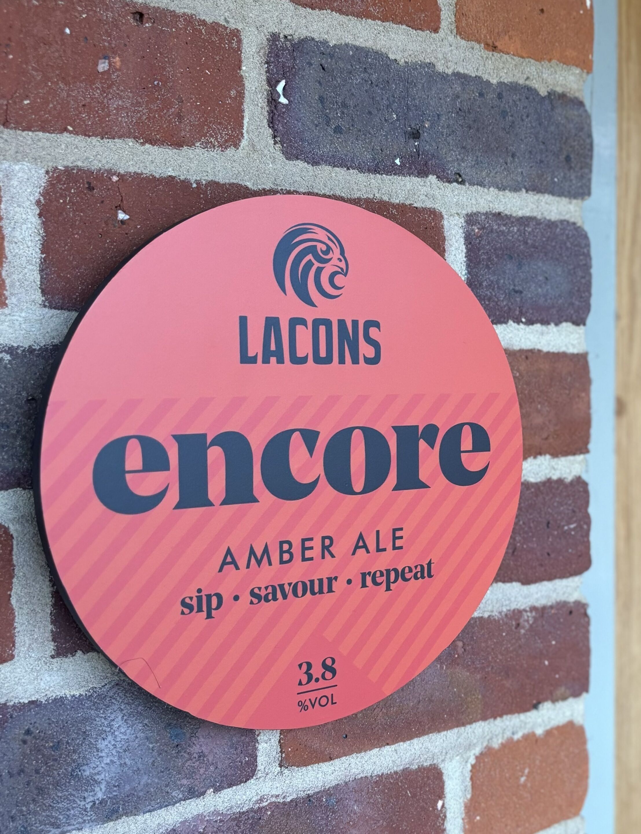 A circular sign with the text "LACONS encore AMBER ALE sip savour repeat 3.8% VOL" mounted on a brick wall.