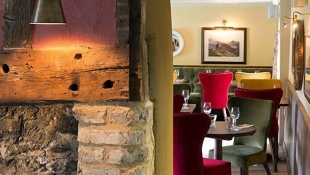 Un intérieur rustique avec des murs en briques apparentes, une poutre en bois et un coin repas agrémenté de chaises à revêtement rouge et d’un canapé vert.