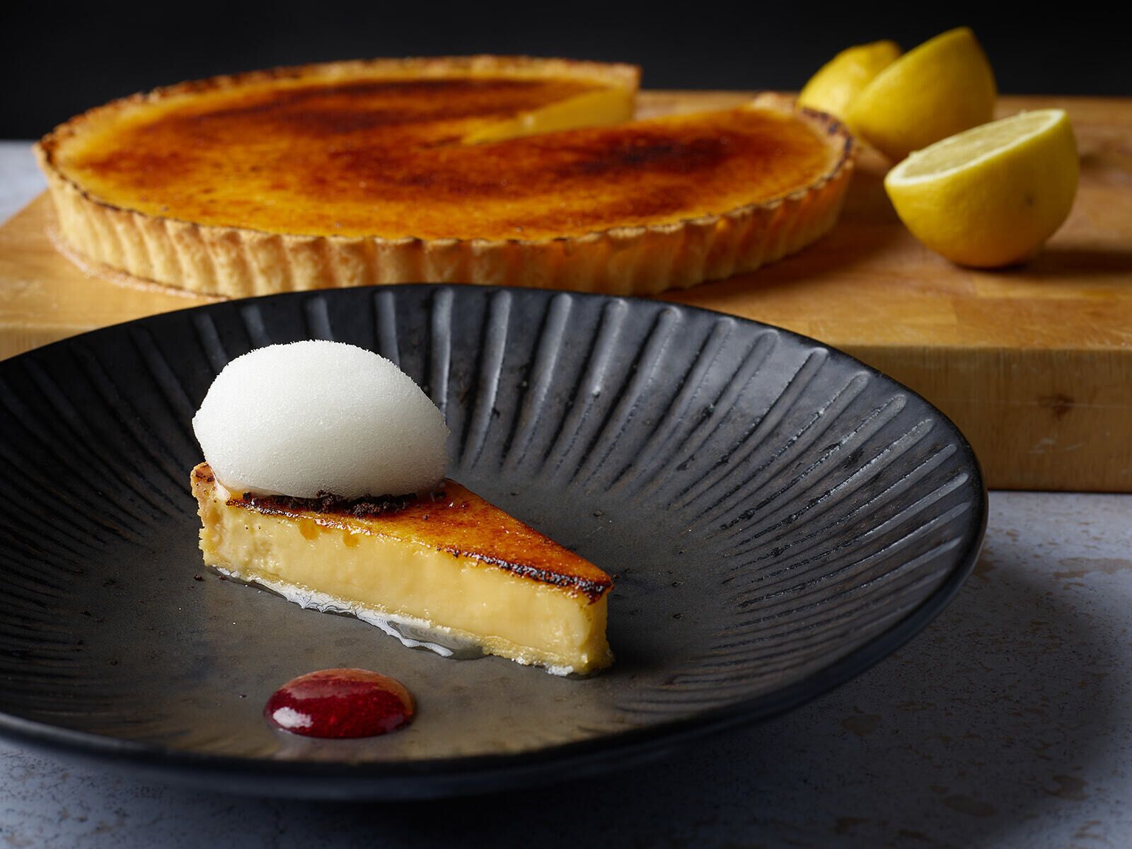 A slice of custard pie with a scoop of ice cream on a plate, accompanied by a whole pie and lemon wedges on a wooden board.