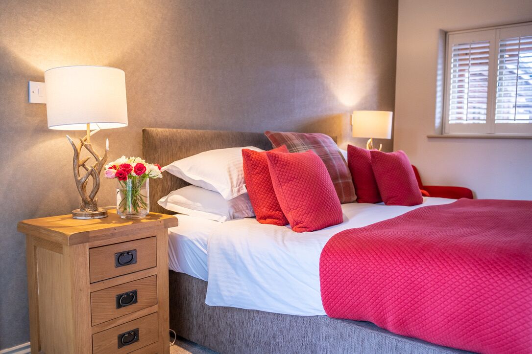 A neatly made bed with red and white bedding, a wooden nightstand with drawers, a vase with flowers, and two wall-mounted lamps.
