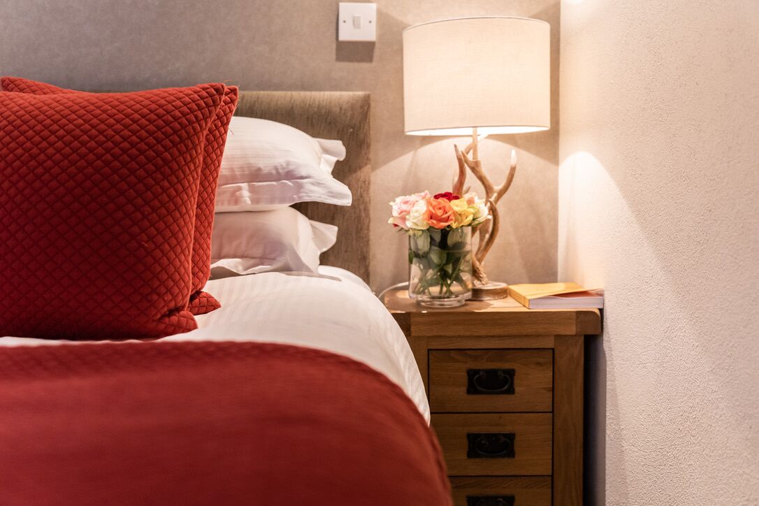 A neatly made bed with a red pillow, a bedside table with a lamp and a vase of flowers, and a wooden nightstand with drawers.