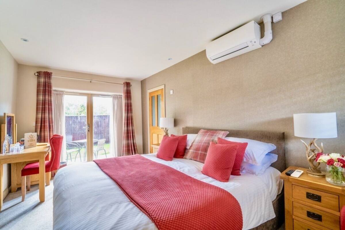 A hotel room with a bed, a desk, and a chair. There is a window with curtains, an air conditioning unit on the wall, and a nightstand with a lamp and a vase of flowers.