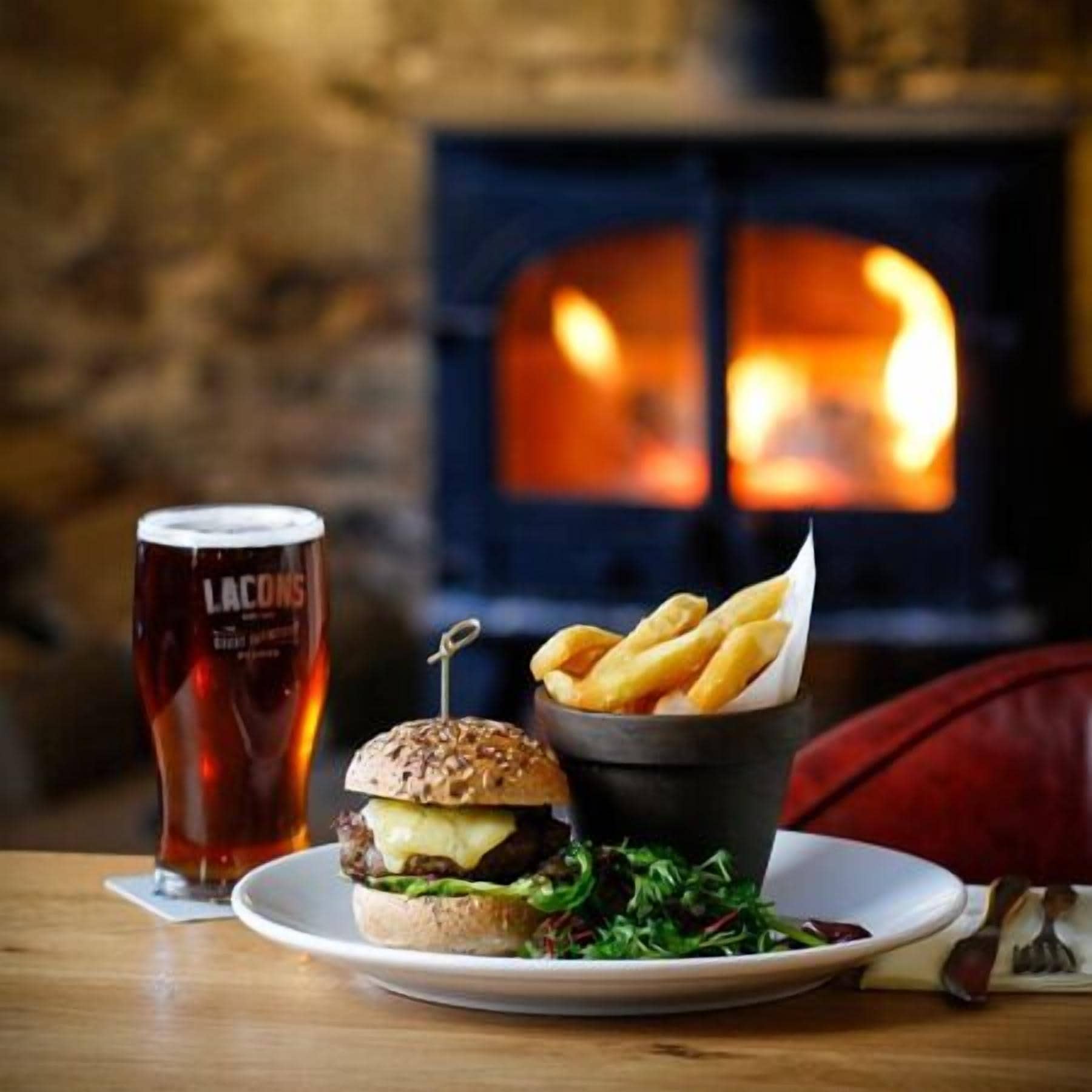 A meal with a burger, fries, and a glass of beer on a wooden table with a fireplace in the background.