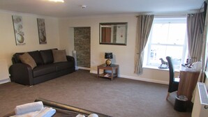A hotel room with a dark brown sofa, a small table with a lamp, a window with curtains, and a view of the outside. - The Southgate Bed And Breakfast (Filey)