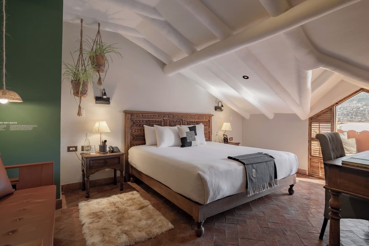 A bedroom with a large bed, two bedside tables, a hanging plant, and a view through a window.